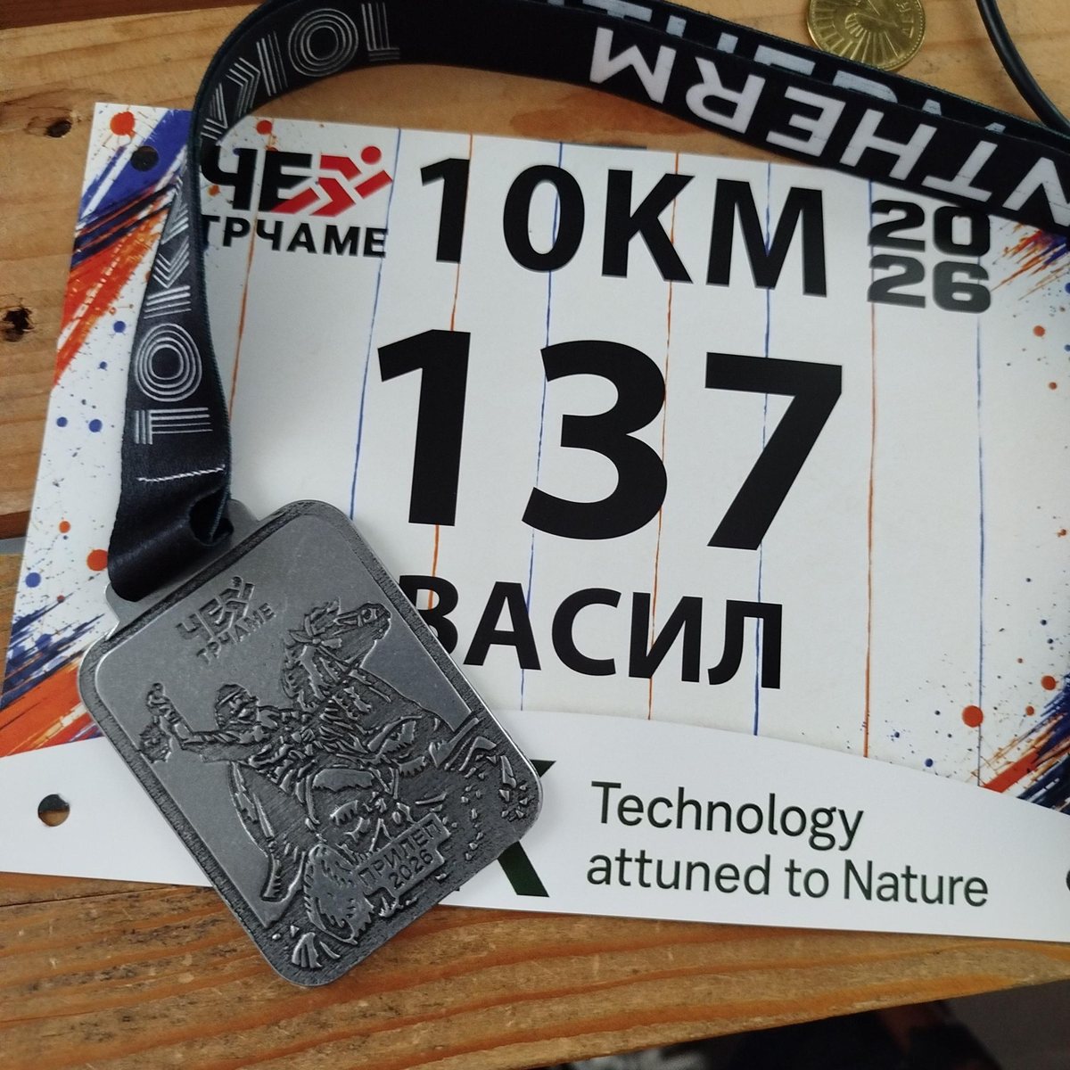 Bib number 137, Васил, 10 KM, Че Трчаме 2026, lying on a wooden table with the race medal on its lanyard across it.
